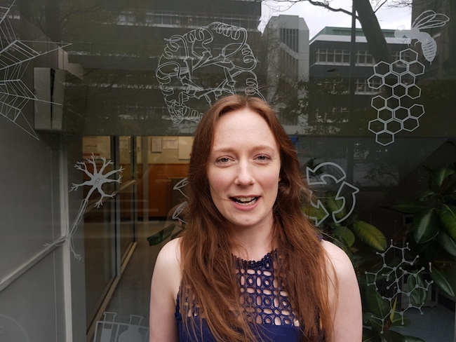 Steph McGimpsey stands outside the Otago Biochemistry Department, explaining her research.