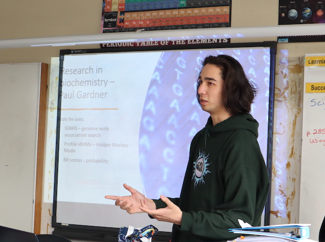 A male university student stands in front of a whiteboard in a class room, talking to an unseen audience.