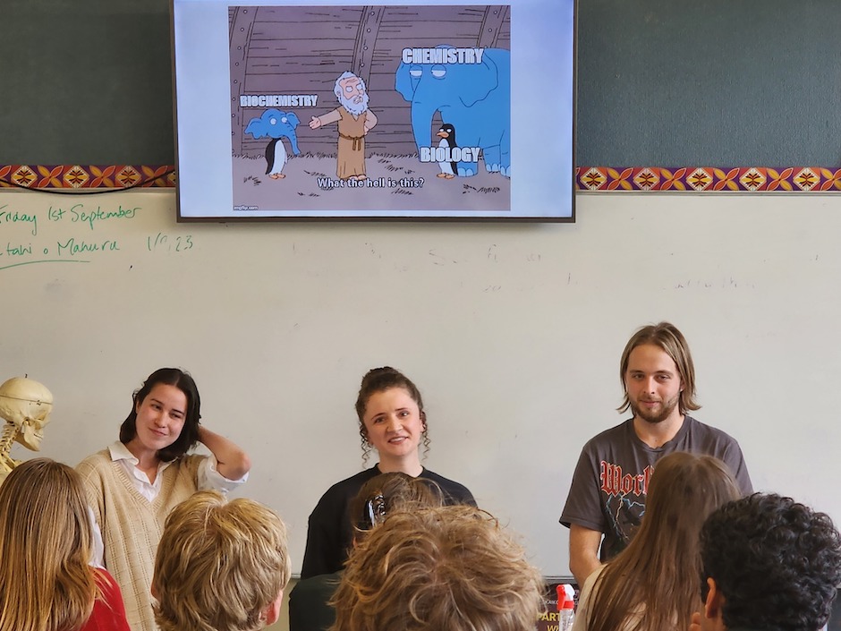 Three students stand at the front of a classroom.