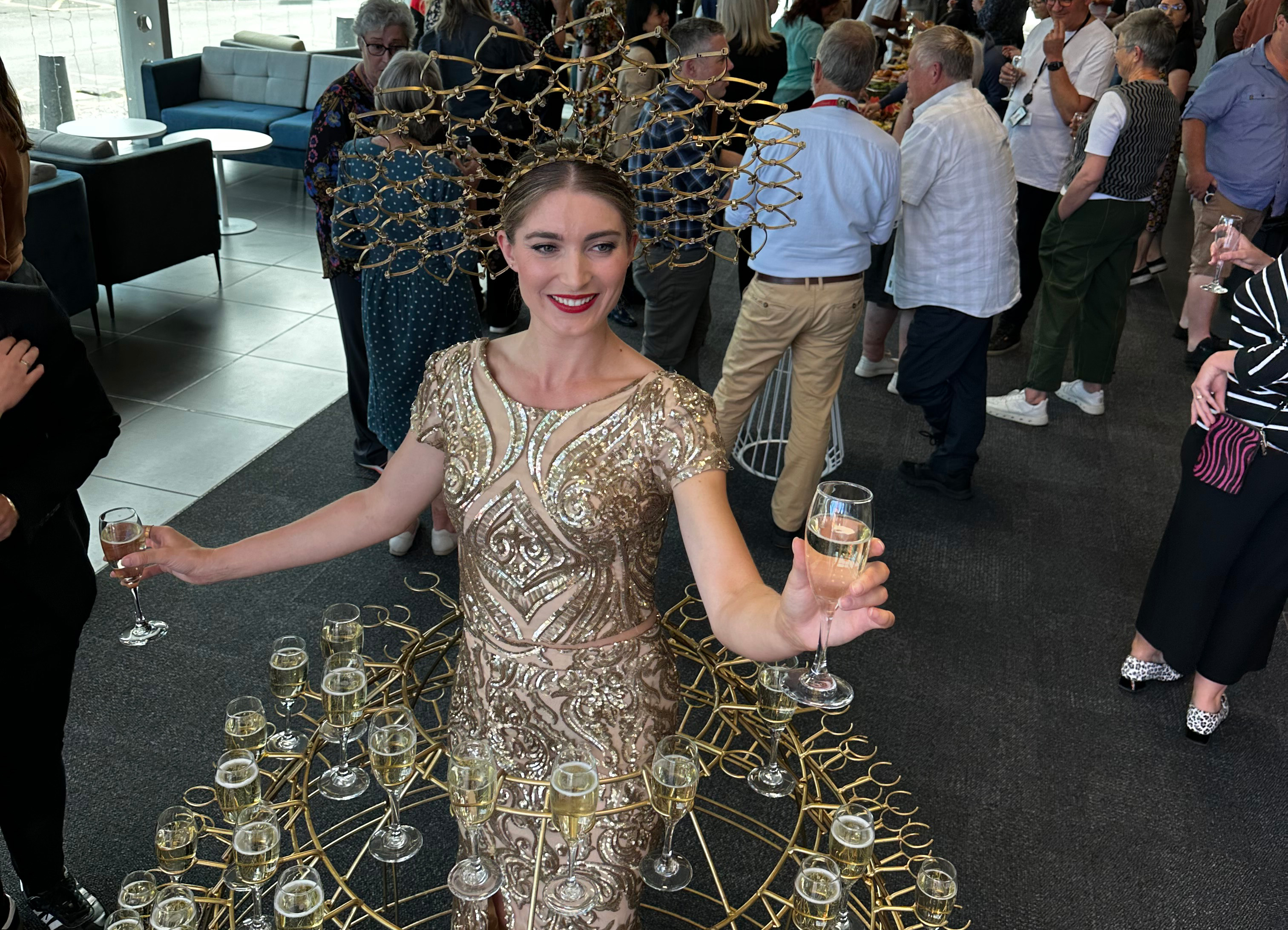 A woman in a gold dress with a hoop skirt that holds many glasses of champagne, with party attendees in the background.