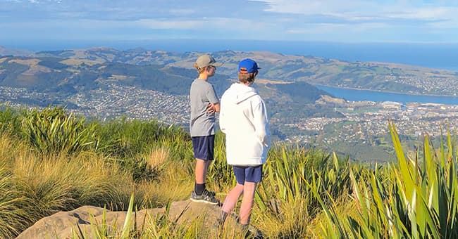 Two students standing on top of a hill surrounded by green flax and tussock, looking over Dunedin city and the Pacific Ocean