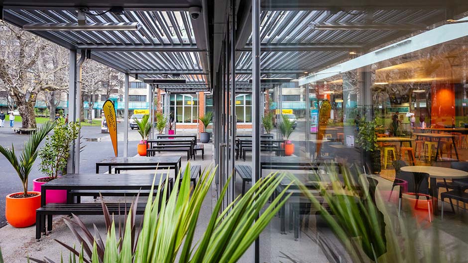 A view of the covered outdoor area of Auahi Ora café, with tables, benches and pot plants sitting outside large glass doors and windows