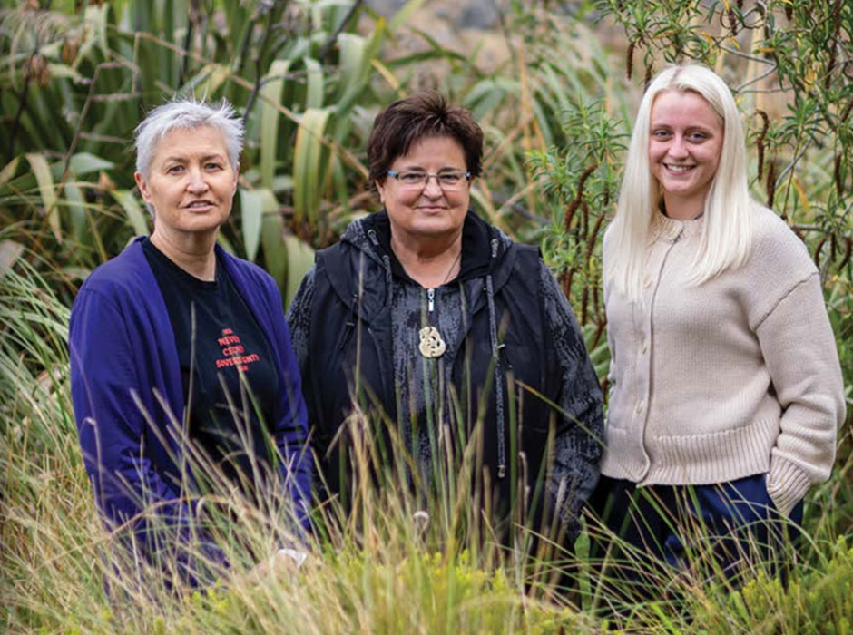 three woman standing behind some tussocks and in front of some flax bushes