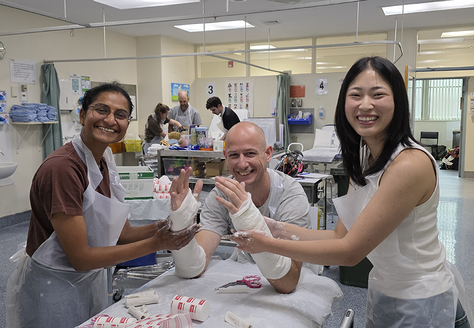 Two female students practise casting the arms of a male patient in a hospital setting.