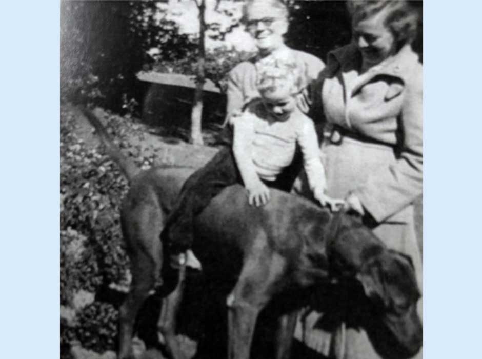 A black and white image of a young girl sitting on top of a dog with a lady standing behind them and another lady standing next to them