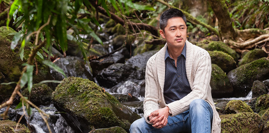 Joon Kim sitting on a rock in a forest next to a stream