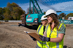 Two students in high-visibilty vests and safety helmets on a building site discussing paperwork