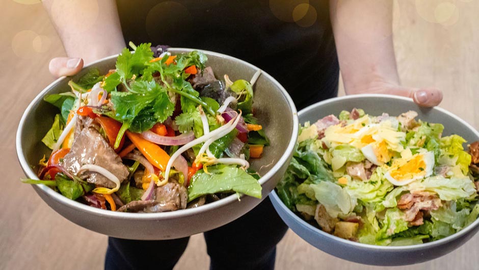 Two bowls of Thai beef and Caesar salads held by an Auahi Ora café staff member