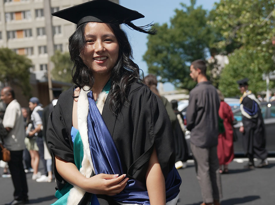 A woman standing in graduation attire on a sunny day with other people standing in the background