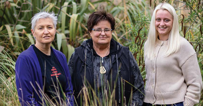 three woman standing behind some tussocks and in front of some flax bushes