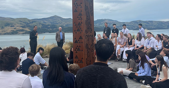 a man speaks, surrounded by young people, on the edge of Akaroa Harbour