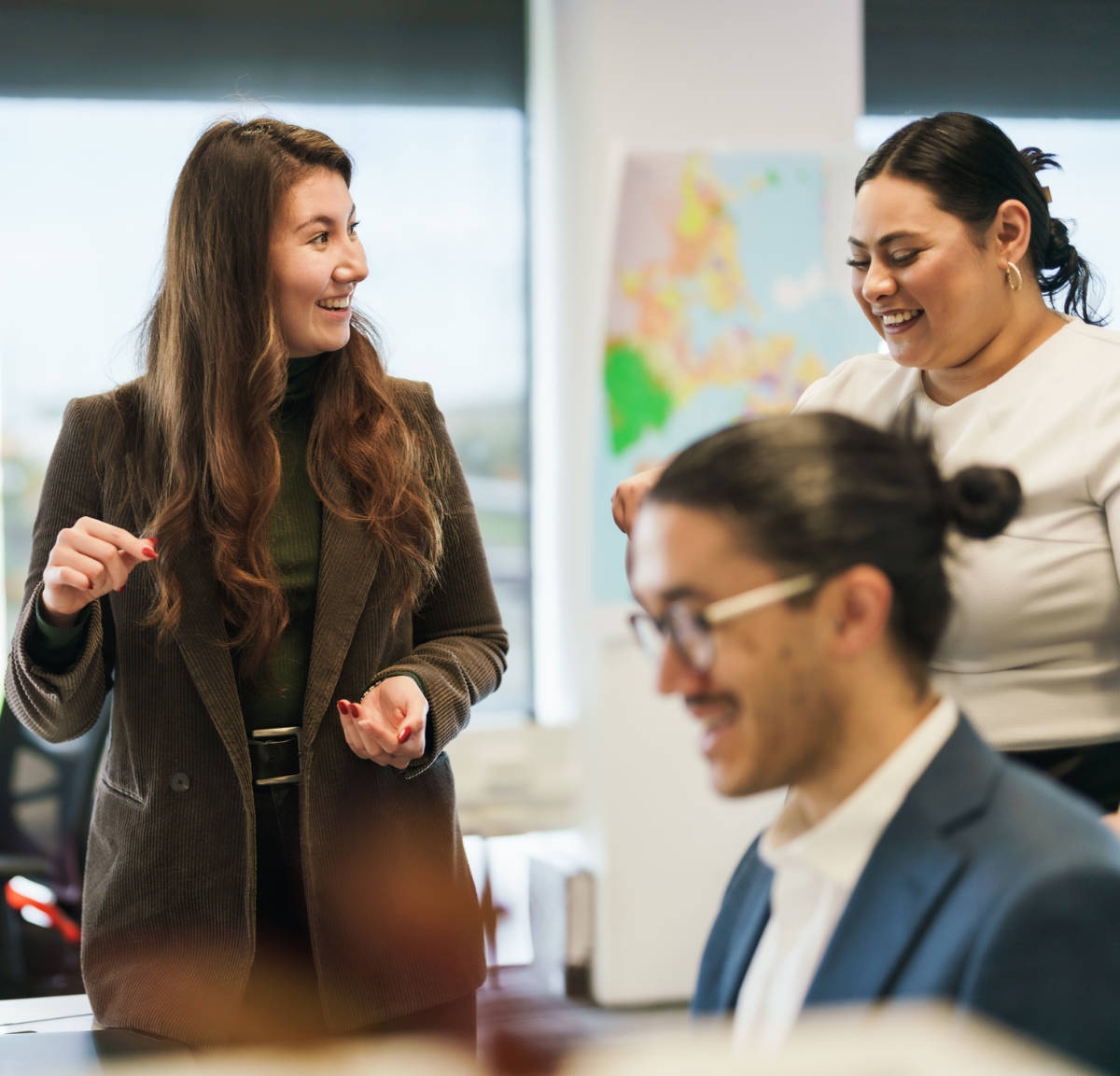 Two women in business attire smiling and talking in a modern office, with a fellow attendee seated in the foreground