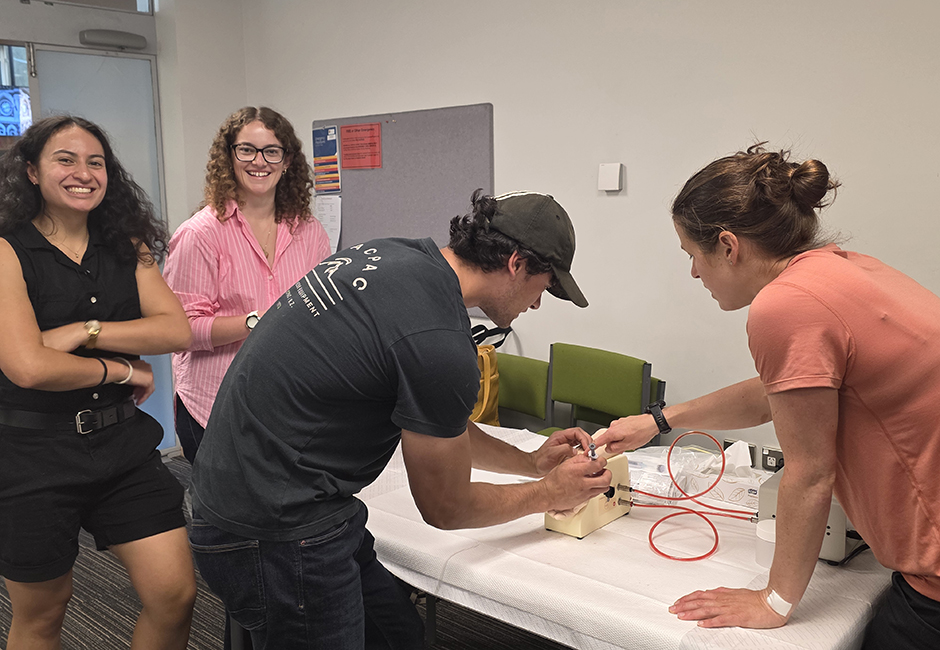 A male and female student practise using medical equimpent on a table while two other female students watch.