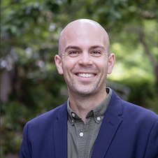 A headshot of a bald, smiling man in front of green trees.