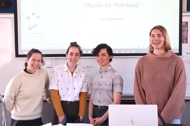 Four young people standing in a line in front of a whiteboard and projector screen, smiling at the camera