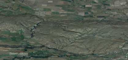 Oblique view of the Raggedy Range from the northwest, with Manuherikia River in foreground and Poolburn valley in background. The smooth gentle slopes of the range reflect the folded structure of the schist bedrock, and this fold is still developing as the range rises. The rising range has forced the Poolburn to cut a deep gorge through the range (left), through which the rail trail passes.