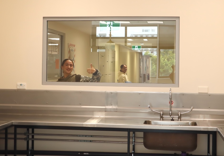 A sink bench with an internal window above it through which can be seen two smiling people, one of which is giving a thumbs up signal.
