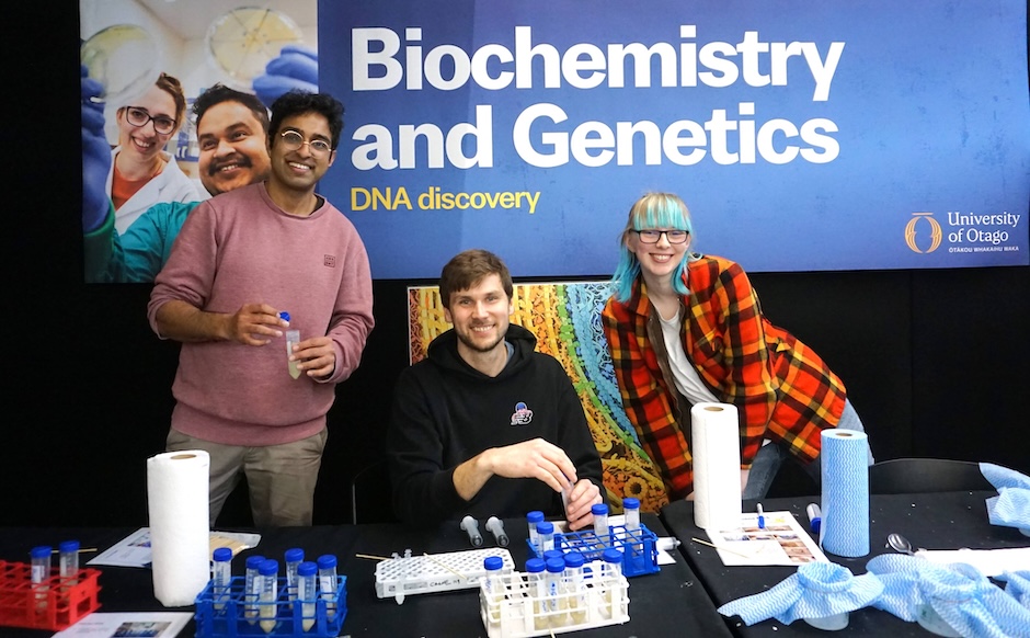 Three smiling people sit and stand behind a desk covered in tubes and racks, a big banner behind them that reads ‘Biochemistry and Genetics: DNA discovery’.