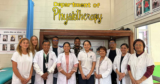 Two rows of people standing underneath a sign over a doorway that says department of physiotherapy, some of the people are wearing white coats