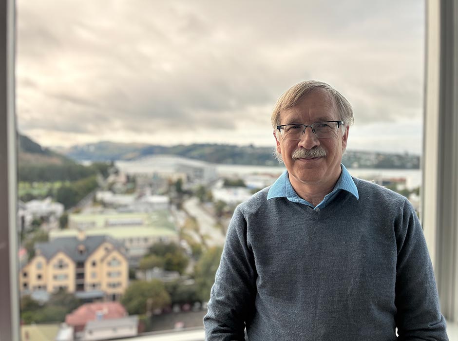 A man standing near a window with a view of the Otago peninsula