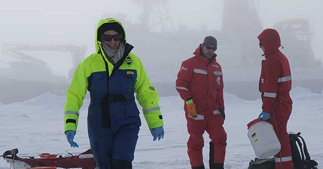 Three people wearing snowsuits standing in snow in Antarctica with a ship in the background foggy