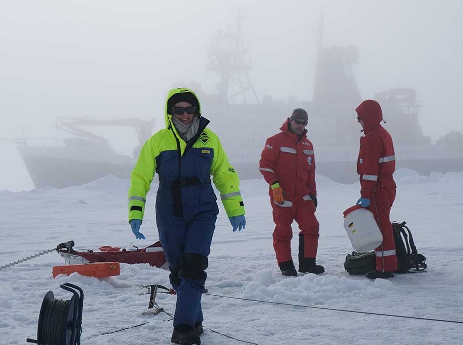 Three people wearing snowsuits standing in snow in Antarctica with a ship in the background foggy