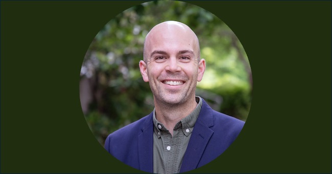 A headshot of a bald, smiling man in front of green trees.