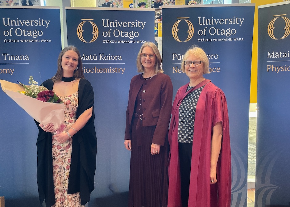 Three women, two wearing academic regalia, stand in front of blue University of Otago banners.