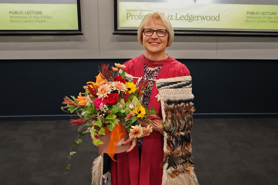 A person in academic dress holds a bouquet of flowers and a ceremonial cloak in a lecture theatre, with screens behind showing “Professor Liz Ledgerwood” and lecture details.