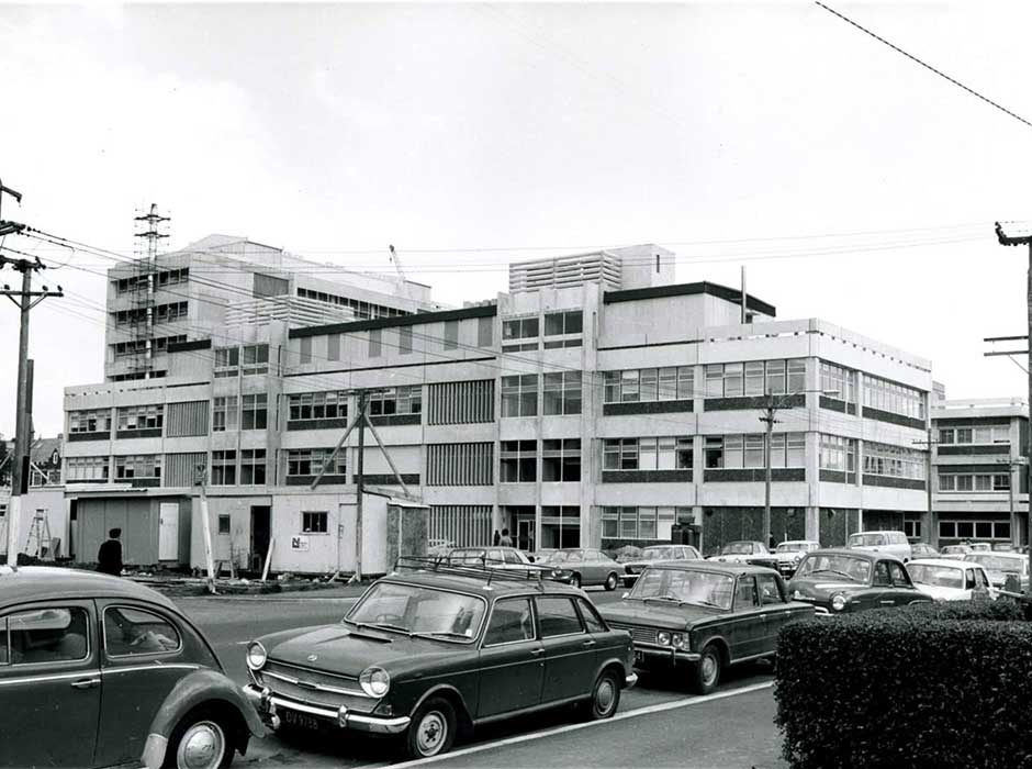 A black and white photo taken in the 19070 with a newly constructed multi-story building in the background and cars in the foreround