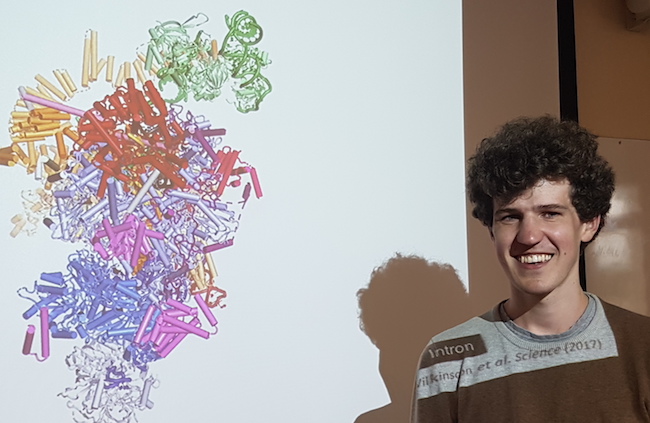 Max Wilkinson standing in front of a picture of a spliceosome.