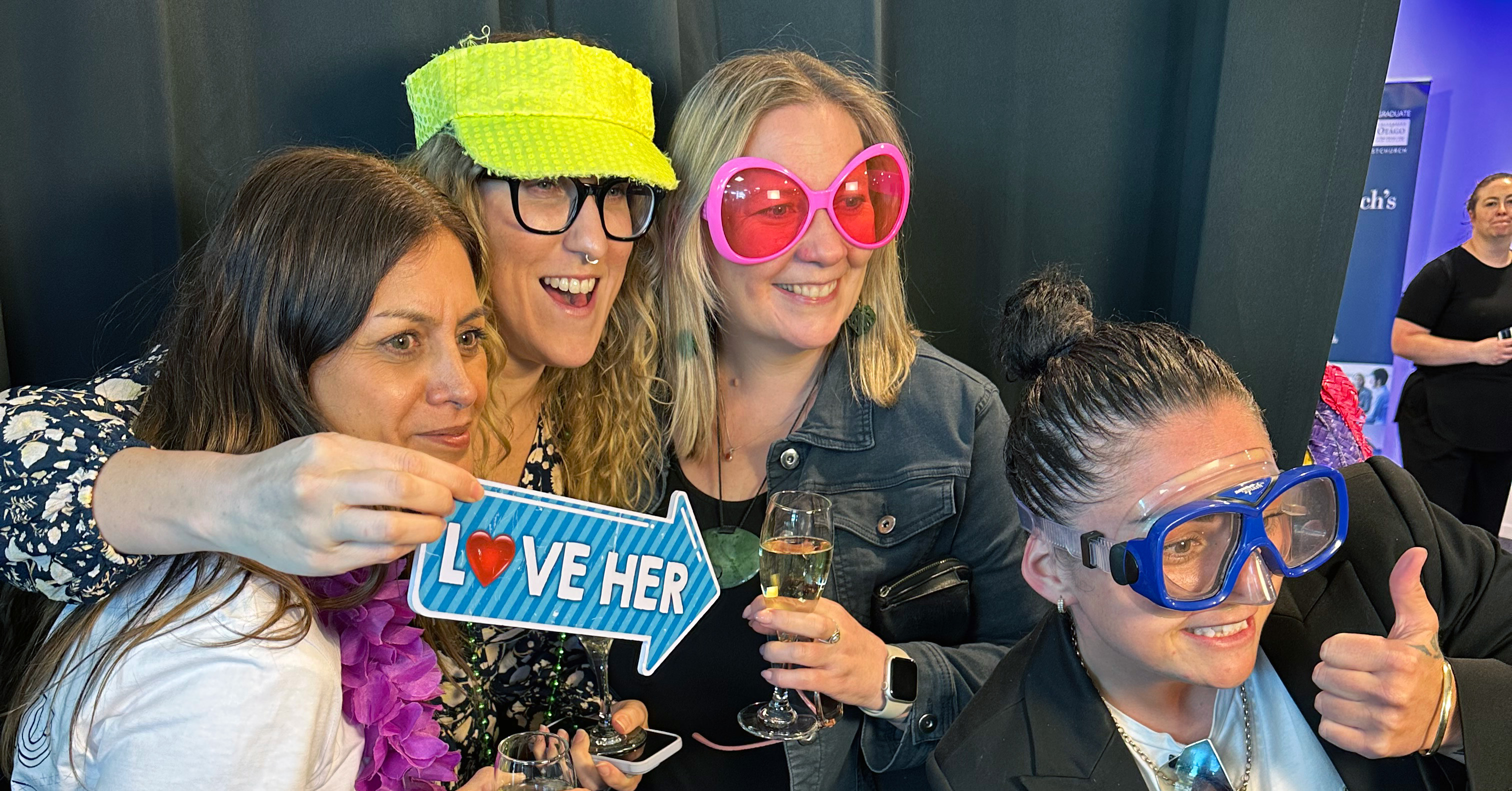 Four women pose for a photo with props including goggles, oversized sunglasses.