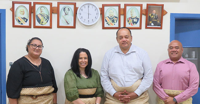 A group of people - two women and two men dressed in traditional Tongan attire, standing in front of a wall with photo frames mounted. 