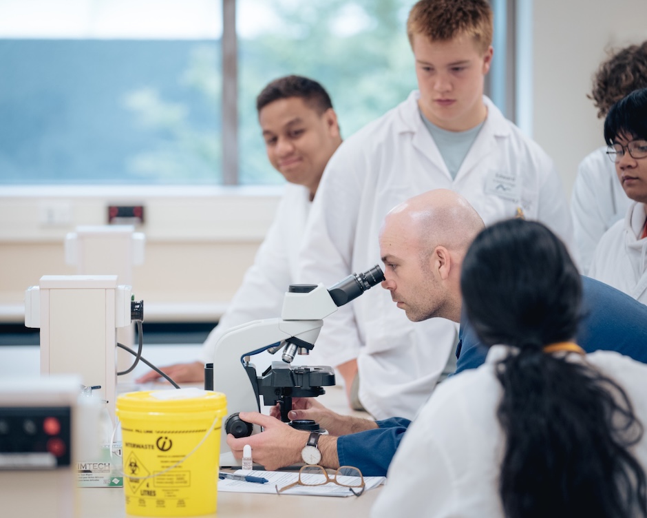 A lab demonstrator in a blue lab coat looks down a microscope while surrounded by five students in white lab coats.