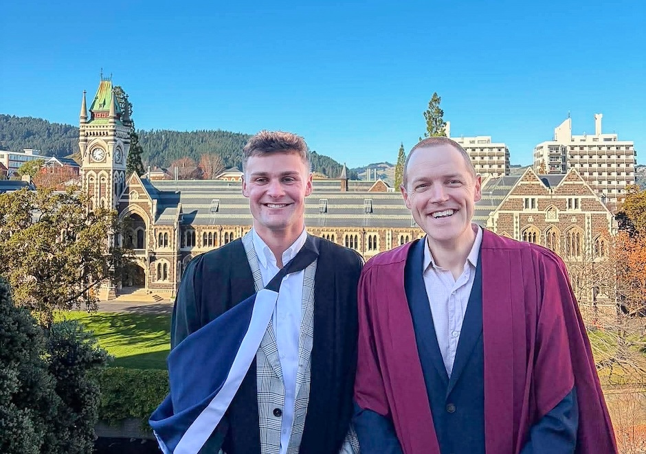 Two men wearing academic regalia stand in front of the University of Otago clocktower buildings.