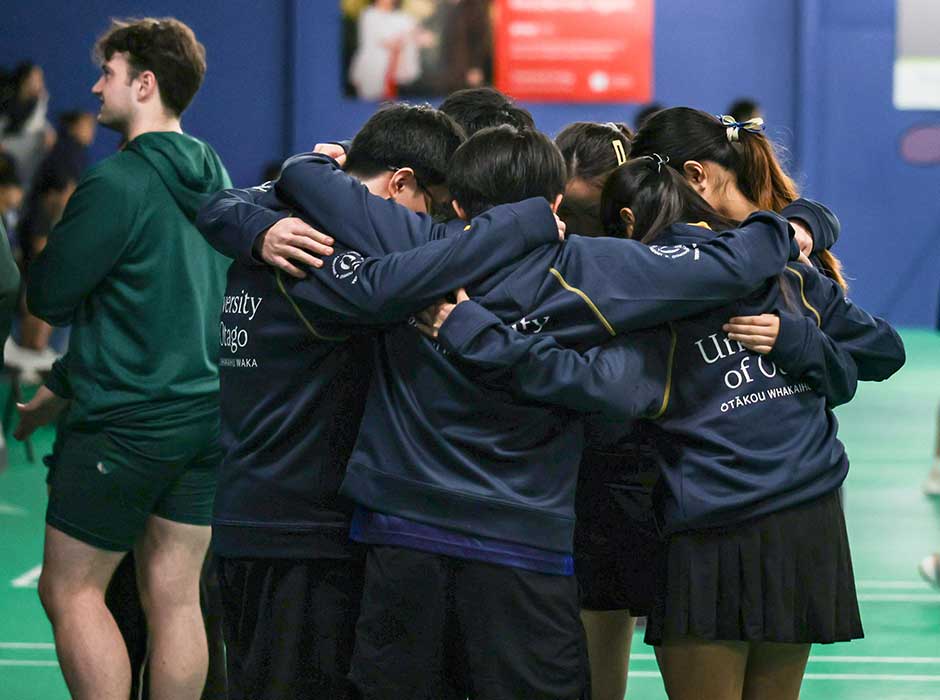 a group of about seven people wearing navy blue Otago hoodies huddled together in a circle