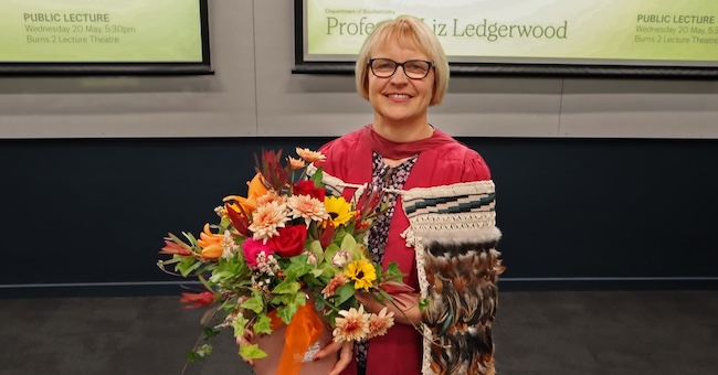 A person in academic dress holds a bouquet of flowers and a ceremonial cloak in a lecture theatre, with screens behind showing “Professor Liz Ledgerwood” and lecture details.