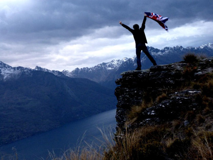 Robin Andrews doing a king of the world pose with nz-flag in arm and NZ remarkables in the background