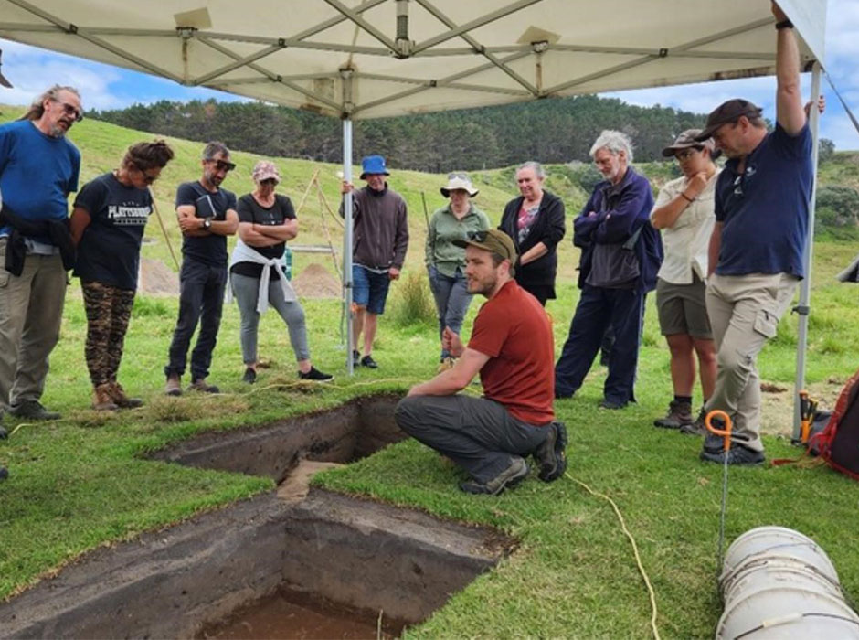 a man crouched down next to a large hole dug in the ground, people standing around him