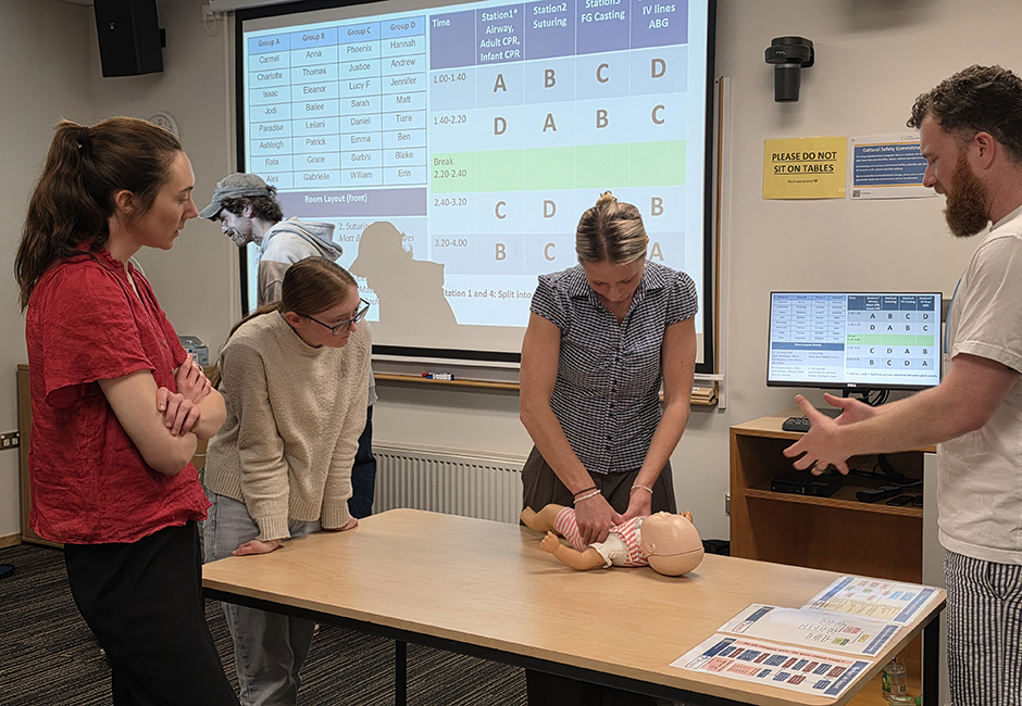 A female student performs CPR on an infant mannequin baby in a classroom setting while two female students and a male tutor watch.