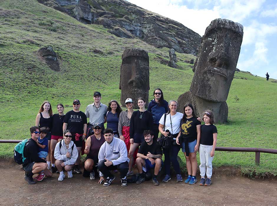 two rows of people all smiling, front row crouched, everyone is in front of a steep, grassy hill with large stone monuments on it