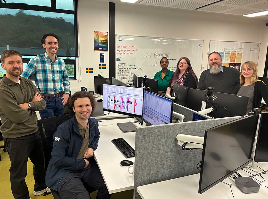 a handful of people sitting and standing around work desks with computers on them