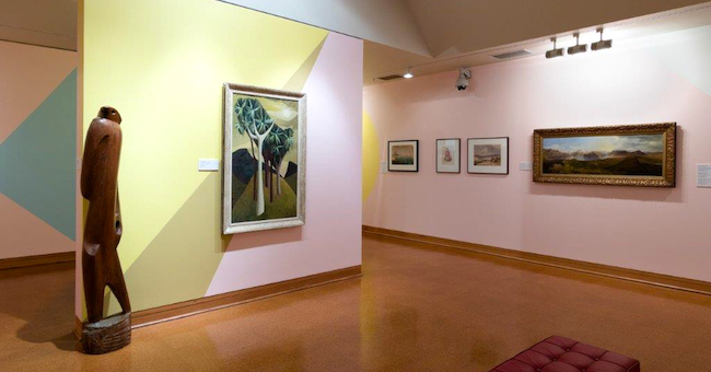 View of a Hocken Gallery exhibition featuring landscape paintings and a large wooden abstract scuplture of a human