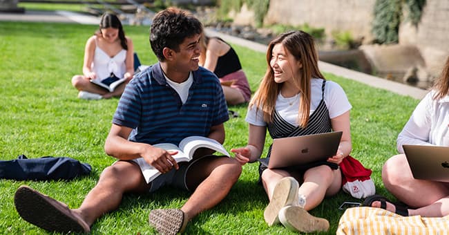 Two smiling students on lawn with laptops and books
