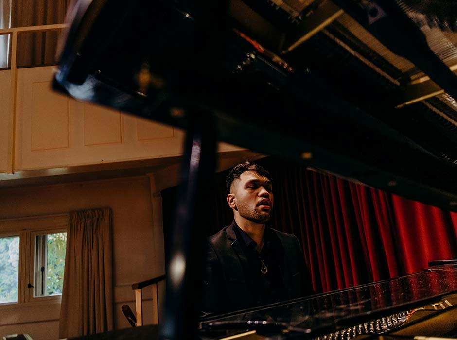 Teone Hotu is pictured sitting at a piano. His passionate about music, and is proud to contribute to the legacy of TRM at Ōtākou Whakaihu Waka.