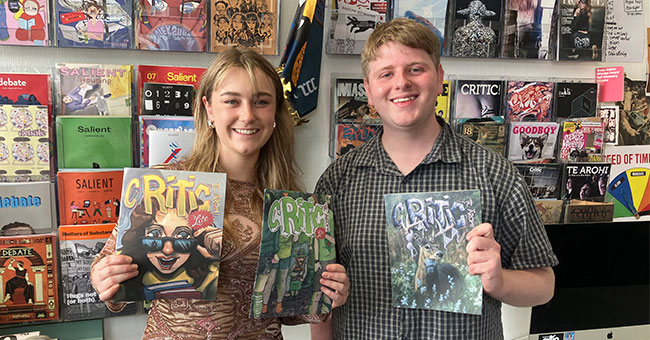 A woman and a man standing next to each other, each holding up a copy of Critic magazine, with many magazines on the wall behind them