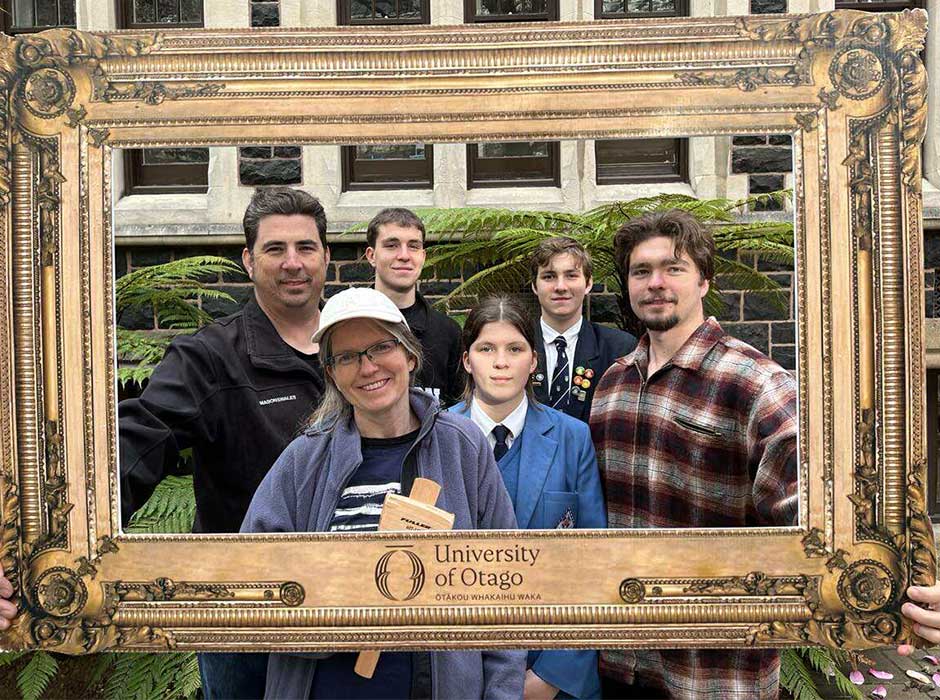 six people standing in front of a stone building and ferns, while holding a large frame up around their head and shoulders