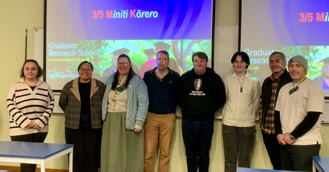 Eight people standing next to each other in front of whiteboards with '3/5 Minit Kōrero' on them