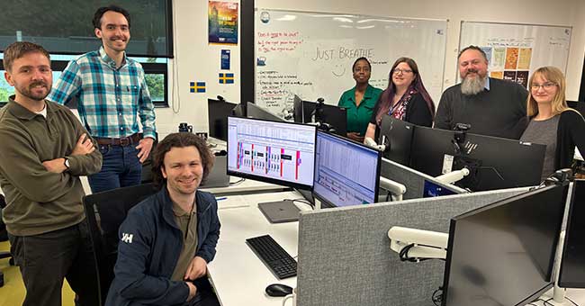 a handful of people sitting and standing around work desks with computers on them