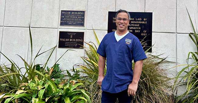 A man wearing blue scrubs standing in front of the faculty of dentistry sign